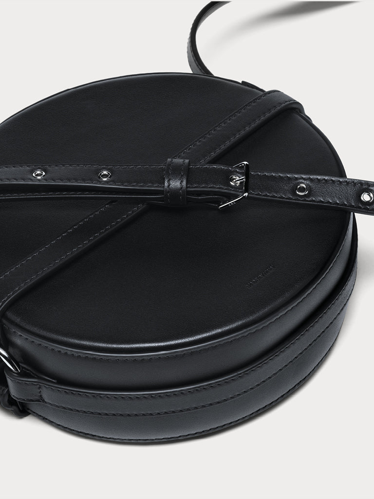 A closeup of a black round crossbody yvy bag with an adjustable strap with silver hardware, horizontal leather band detail, and a smooth leather surface. 