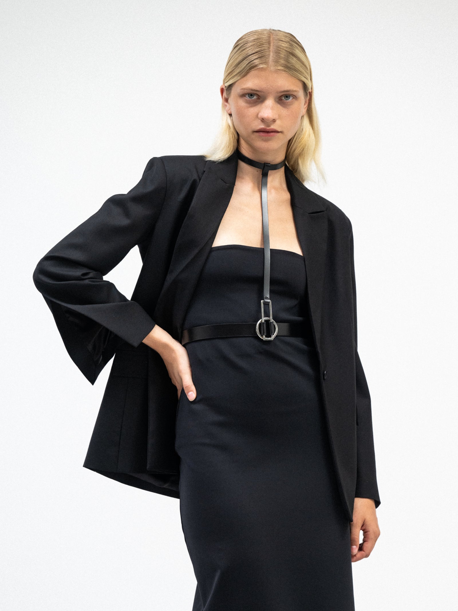A female model wearing the black tube dress with tailored black yvy suit jacket, paired with the statement leather belt with metallic buckle 