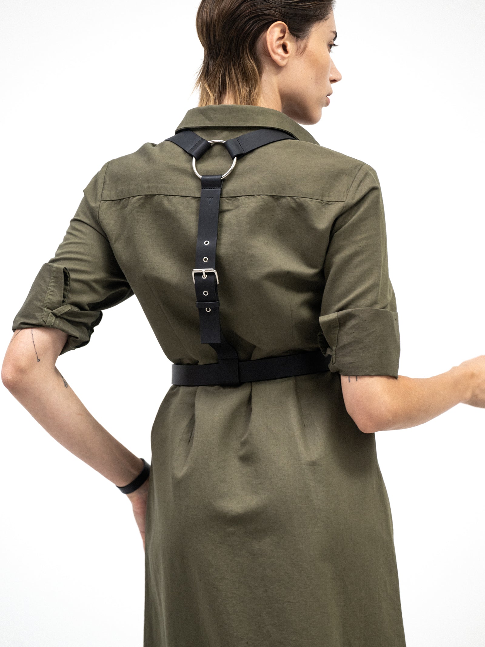 Back view of a woman wearing an olive green shirt dress with rolled-up sleeves, styled with a black harness belt featuring silver hardware. The dress has a relaxed fit and minimalist design.