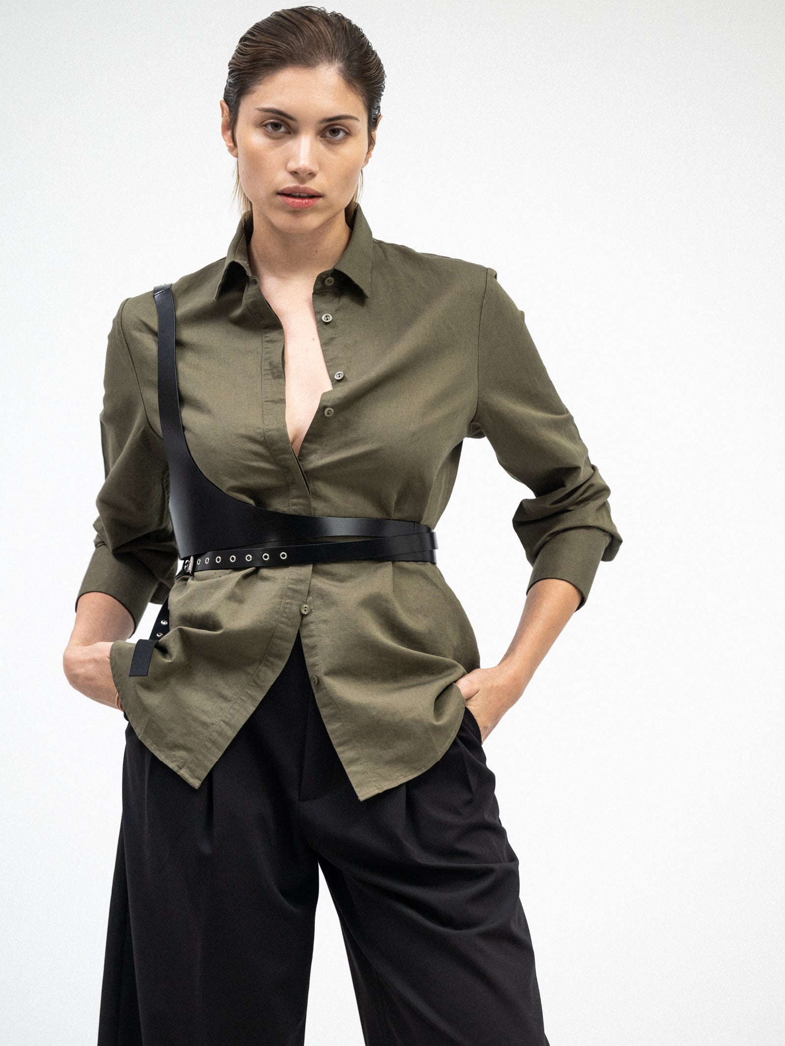 Woman wearing yvy olive green buton-up shirt with a black harness-style belt, paired with loose black trousers. The shirt has a relaxed fit with partially unbuttoned front, creating a modern, edgy look.