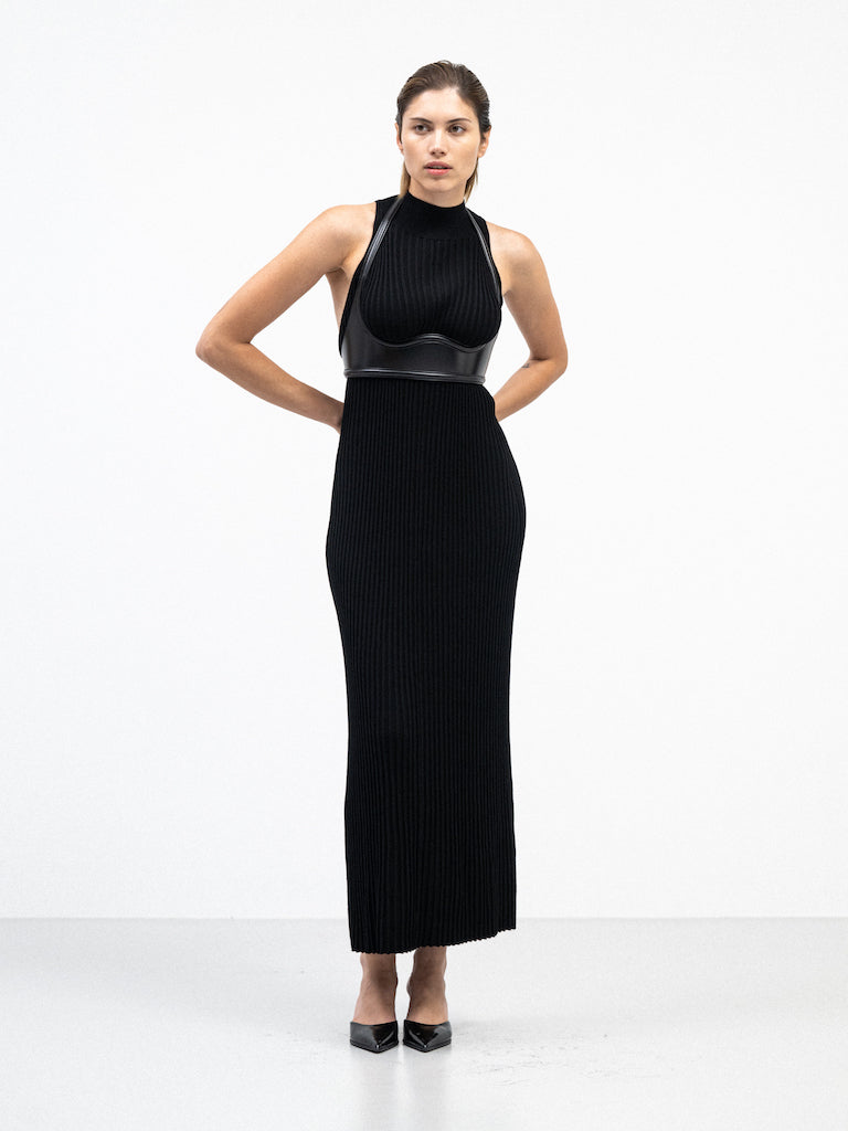 A female model wearing a sleek, black, ribbed yvy dress with a structured leather harness detail, standing in a minimalist white space. She is wearing pointed black heels