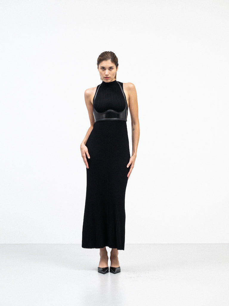 A female model standing in a black ribbed halter-neck dress with a structured leather harness detail, standing in a minimalist white space. She is wearing pointed black heels.