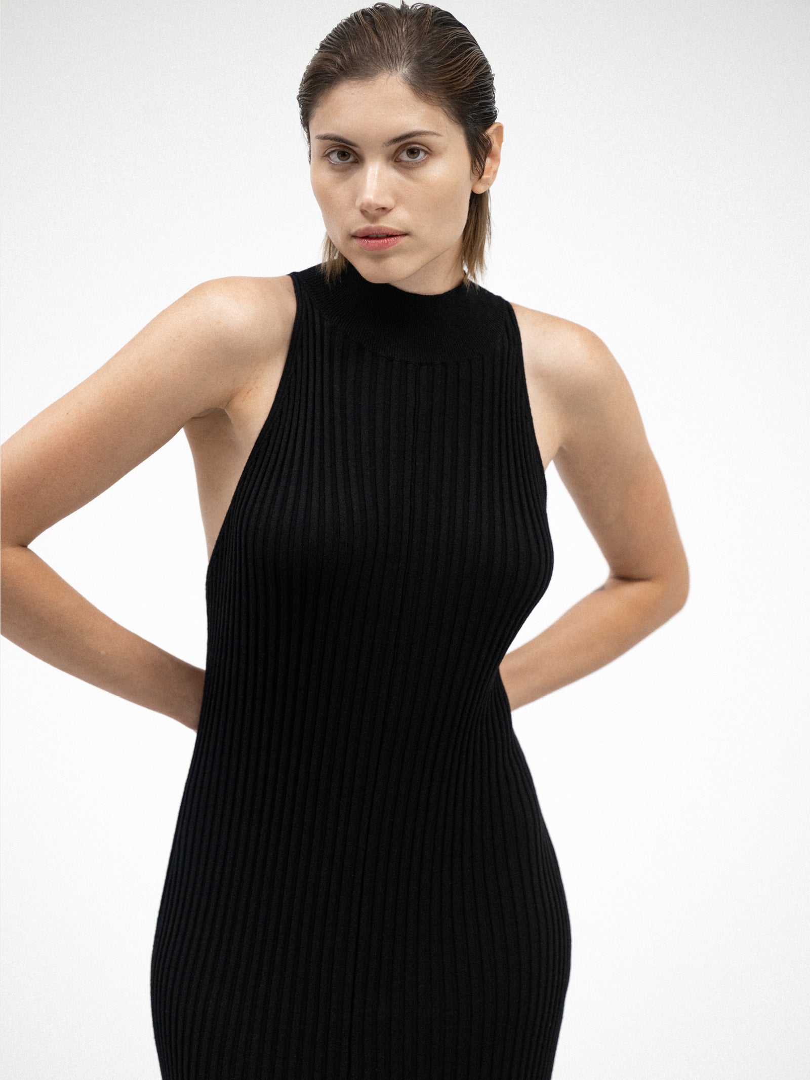 A female model standing in a black ribbed halter-neck dress in a minimalist white space.