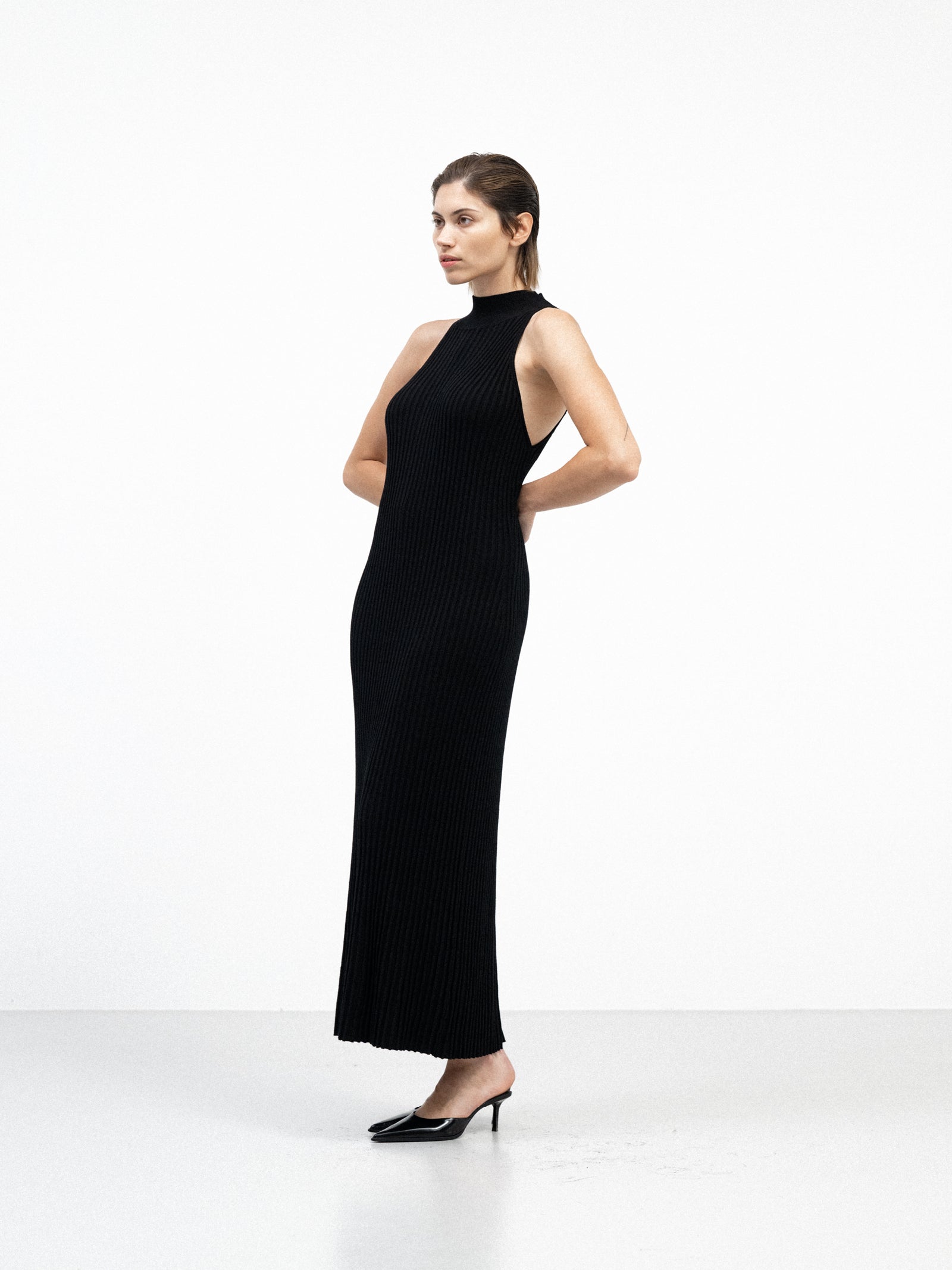 A female model wearing a sleek, black, ribbed yvy dress, standing in a minimalist white space. She is wearing pointed black heels