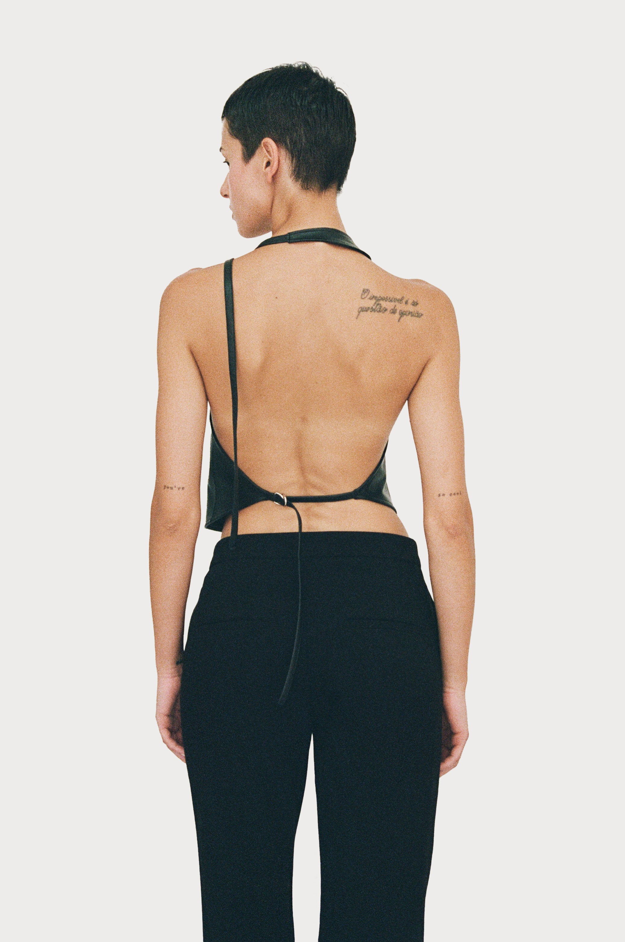 a female model wearing black yvy leather halter neck top with open back and black pants, back view