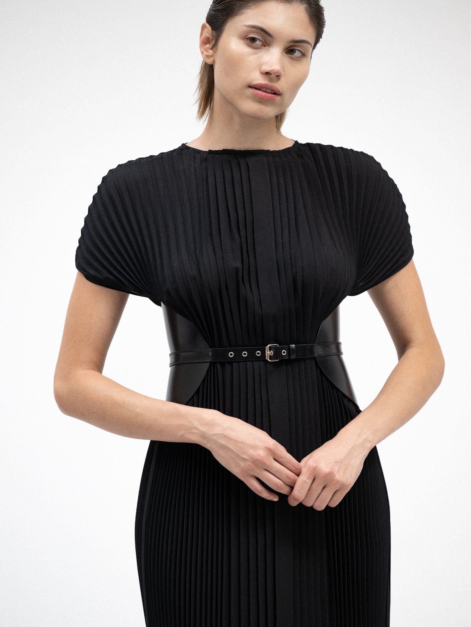A female model wearing black yvy plisse dress cinched with statement black leather corset belt featuring gold hardware
