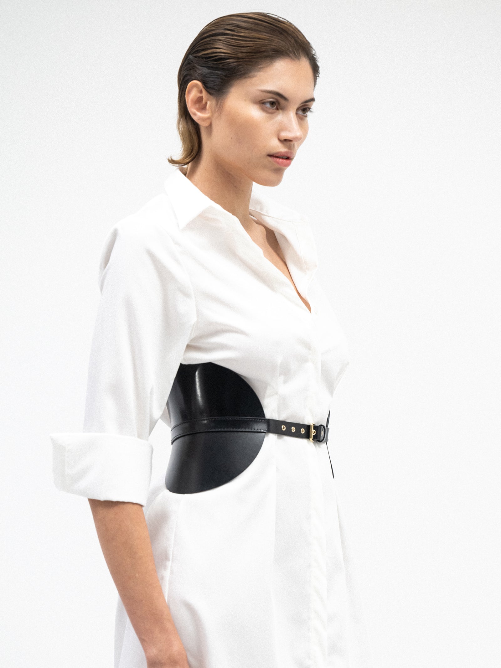 A female model wearing white oversized button-down yvy shirt dress cinched with statement black leather corset belt featuring gold hardware