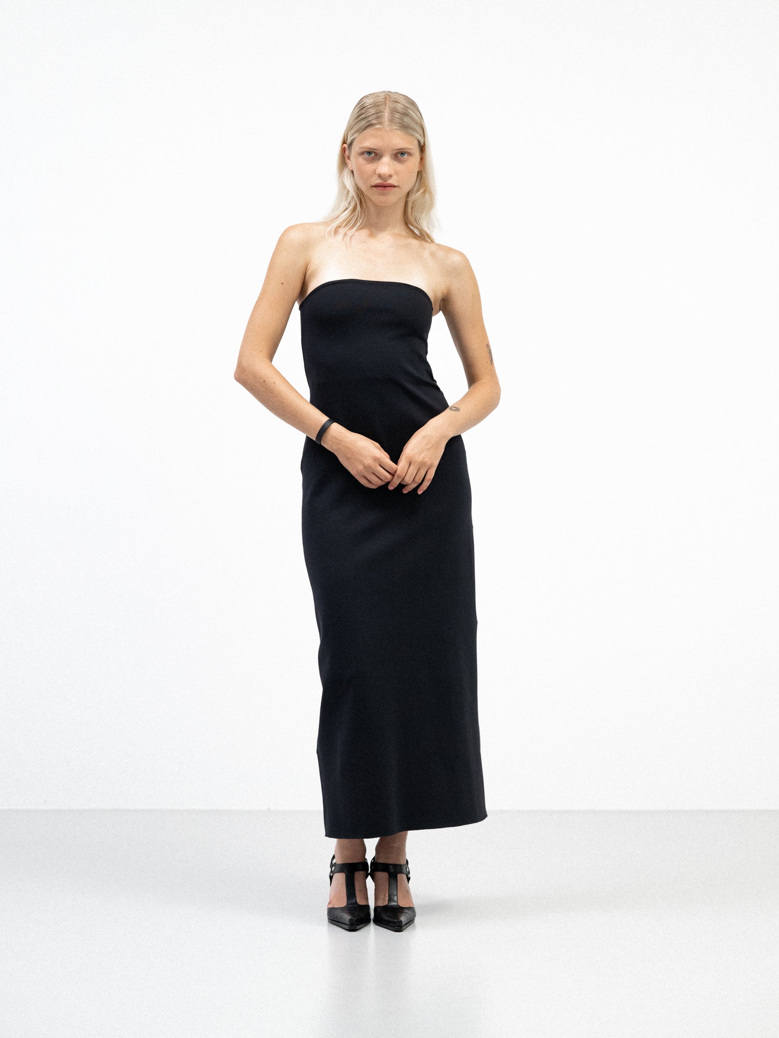 A female model wearing a sleek, black, strapless yvy tube midi dress with a fitted silhouette, standing in a minimalist white space. She is wearing pointed black heels