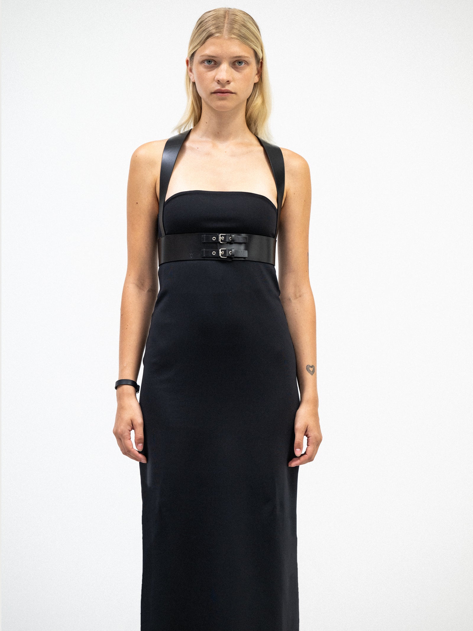 A female model wearing a sleek black tube midi dress styled with a wide black leather harness featuring buckle details, standing in a minimalist white space.