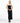 A female model wearing a sleek, black, strapless yvy tube midi dress with a fitted silhouette, standing in a minimalist white space. She is wearing pointed black heels