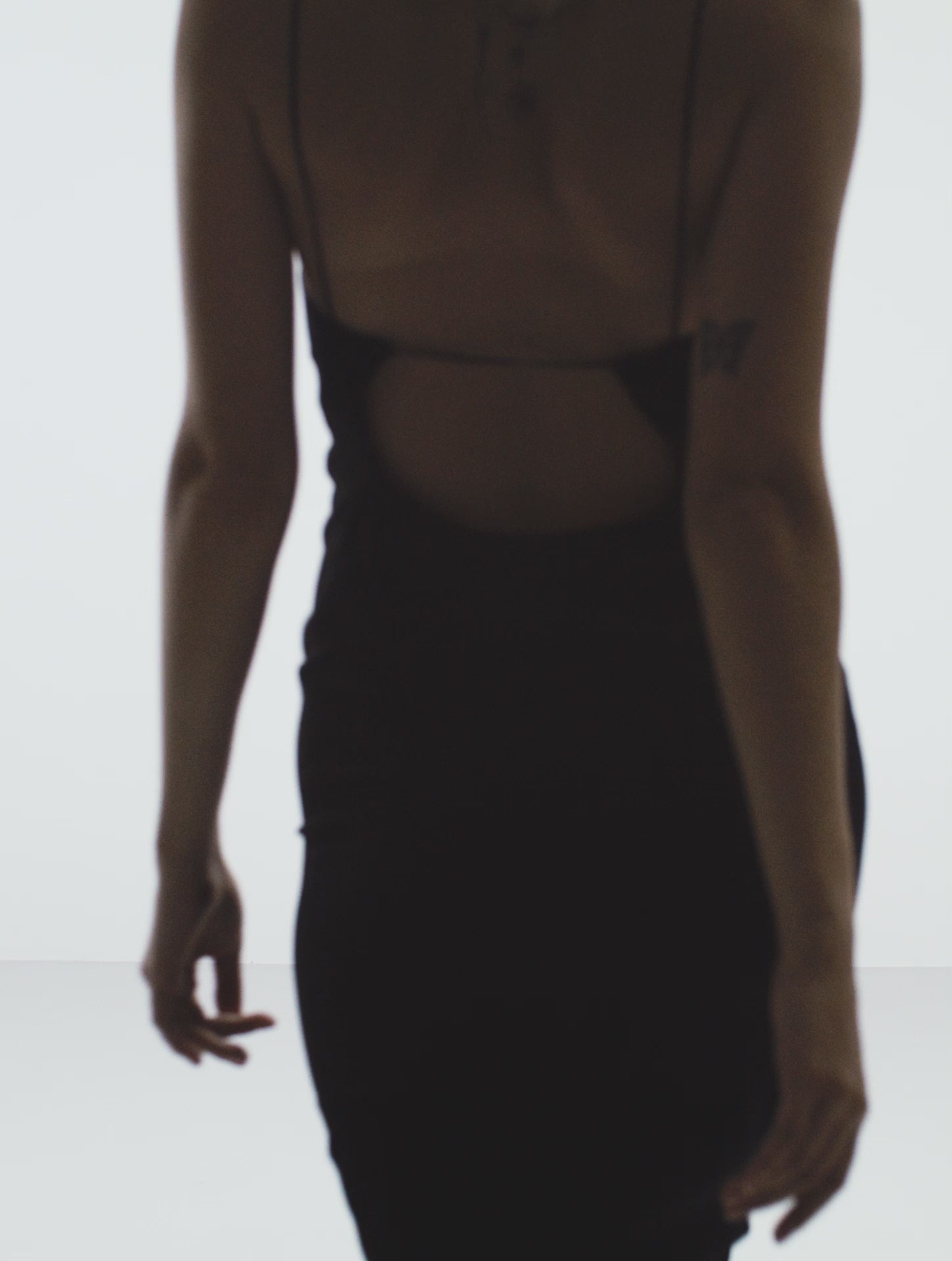 A female model wearing an elegant midi dress that features a curved open back design with plunge back, spaghetti straps, and a form-fitting silhouette