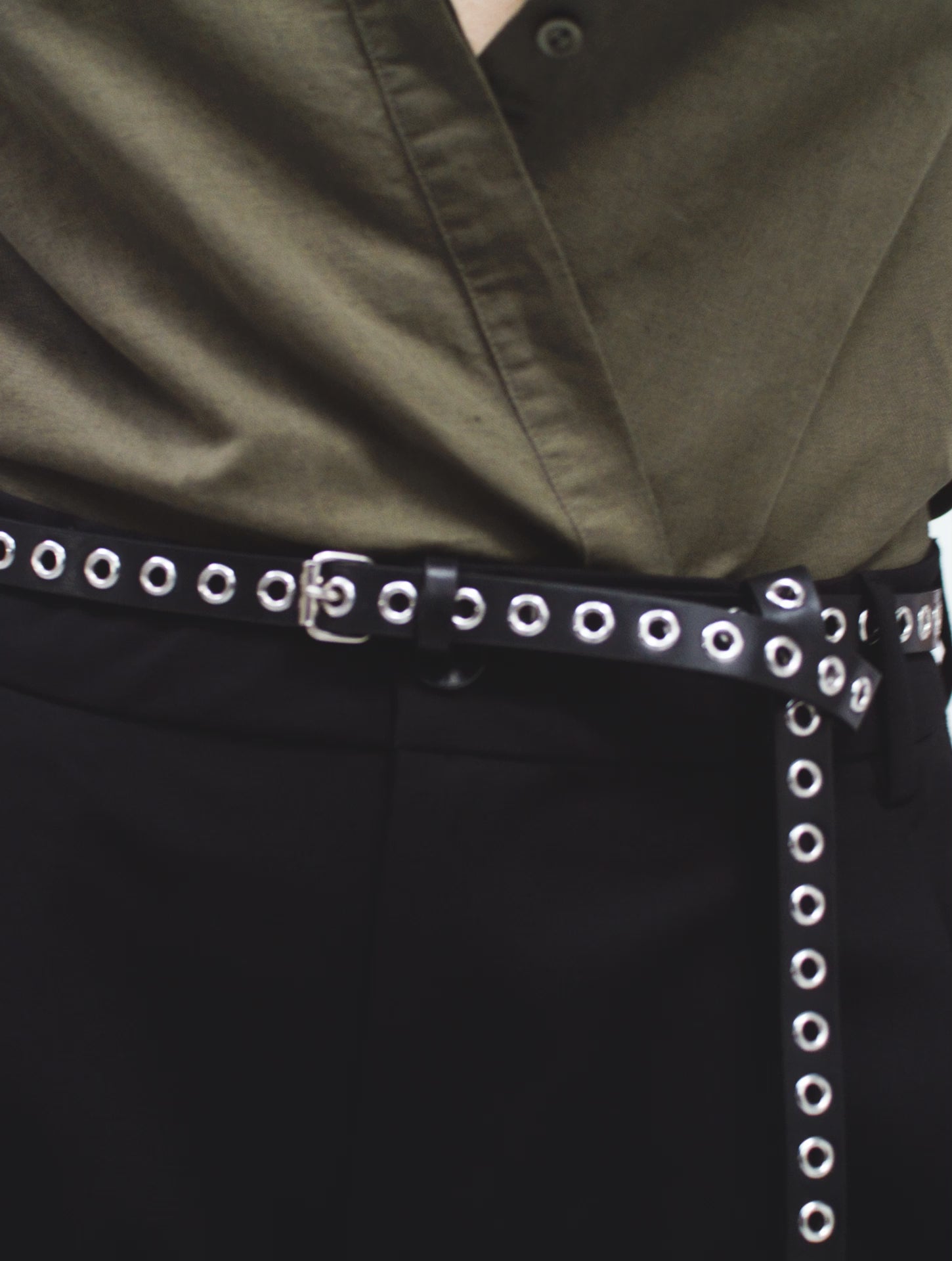 a video of a female model posing in thin black yvy eyelet belt with silver hardware over black pants, yvy leather eyelet choker and green linen long sleeve shirt